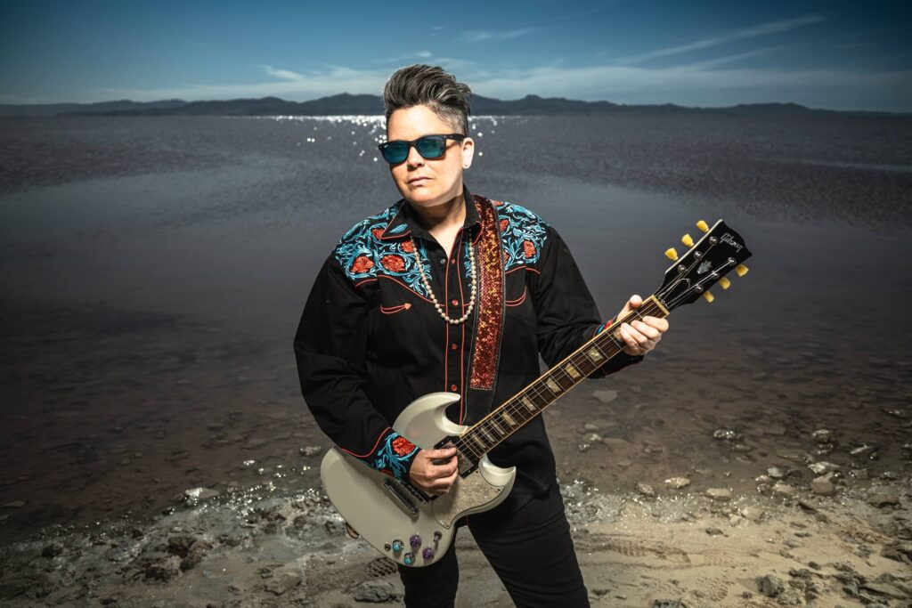 Talia Keys, wearing sunglasses and a black shirt with vibrant floral embroidery, plays an electric guitar while standing on a rocky shoreline. A calm body of water and distant mountains are visible under a clear blue sky.