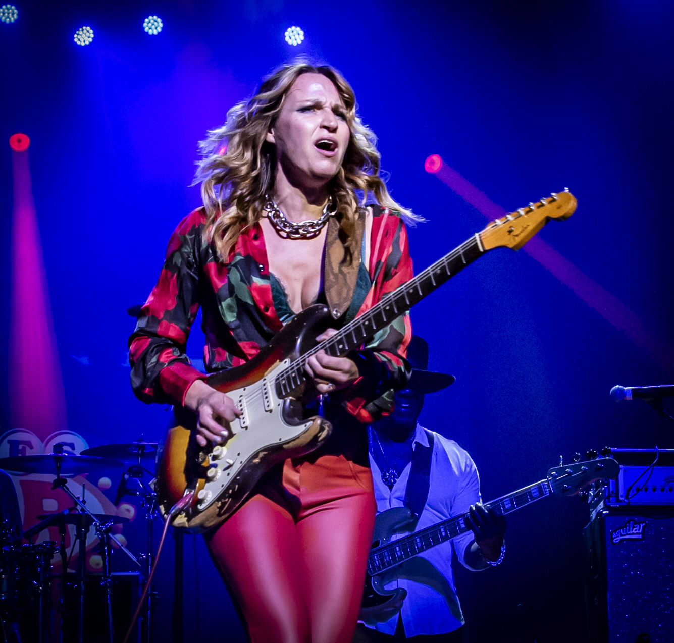 Ana Popovic, wearing a vibrant floral blouse and red pants, passionately plays an electric guitar on stage under dramatic blue and purple lighting. A bassist in the background adds depth to the lively performance, with the stage adorned by glowing spotlights.