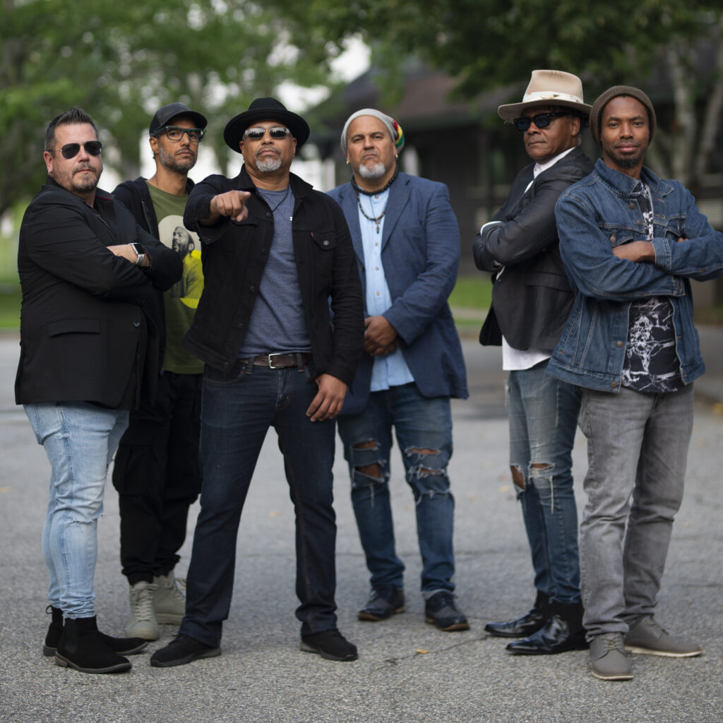 The Blackburn Brothers band, a group of six men, stand confidently outdoors in casual and stylish attire. The members are arranged in a semi-circle, with some crossing their arms and others posing dynamically. The background features greenery and blurred structures, creating a relaxed yet commanding atmosphere.