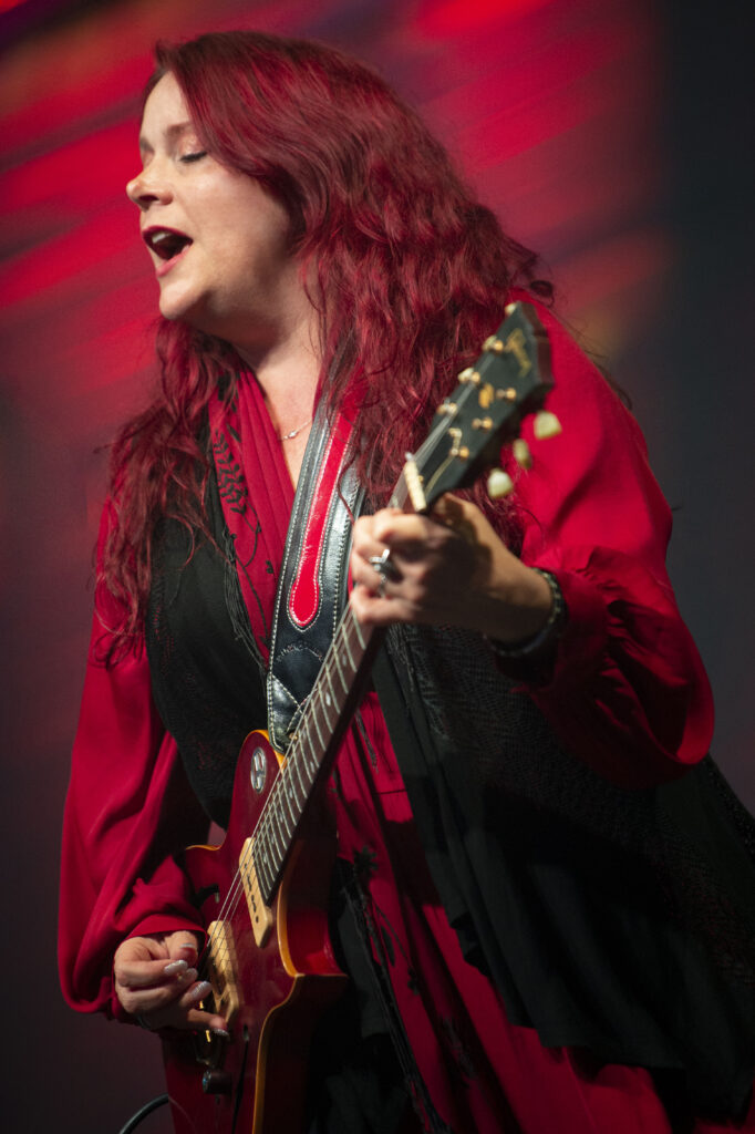 Carolyn Wonderland performing live on stage, singing passionately while playing a red electric guitar. She wears a red and black outfit, with her long red hair flowing under dramatic stage lighting.