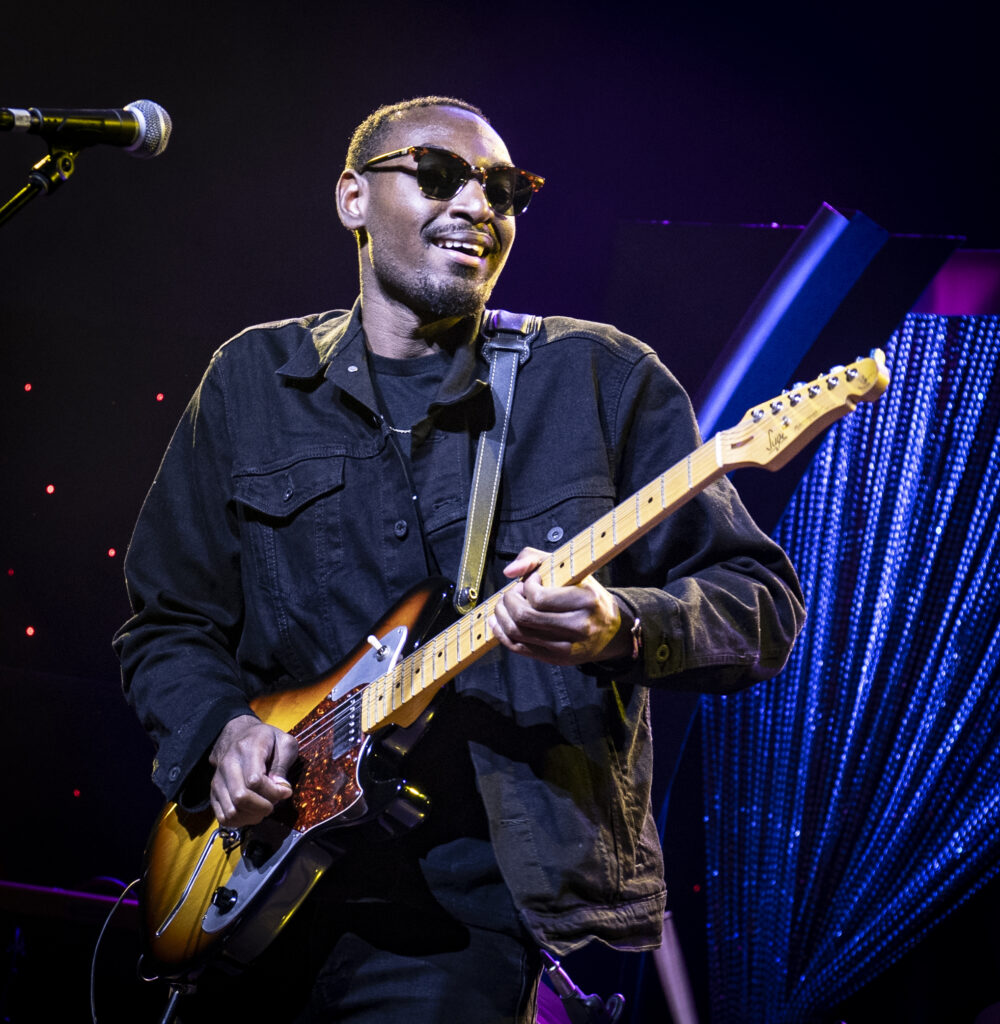 Mathias Lattin, wearing sunglasses and a black jacket, smiles while playing an electric guitar on stage. A microphone is visible to the side, and the background features vibrant stage lighting and decorative elements.