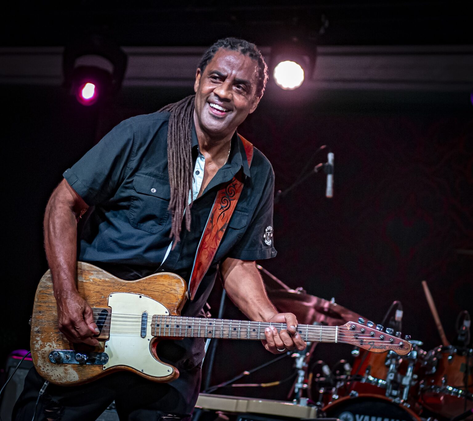 Kenny Neal, a musician with long dreadlocks, smiles while playing a worn-out electric guitar on stage. He is wearing a black shirt and an orange guitar strap, with stage lighting and drums in the background.