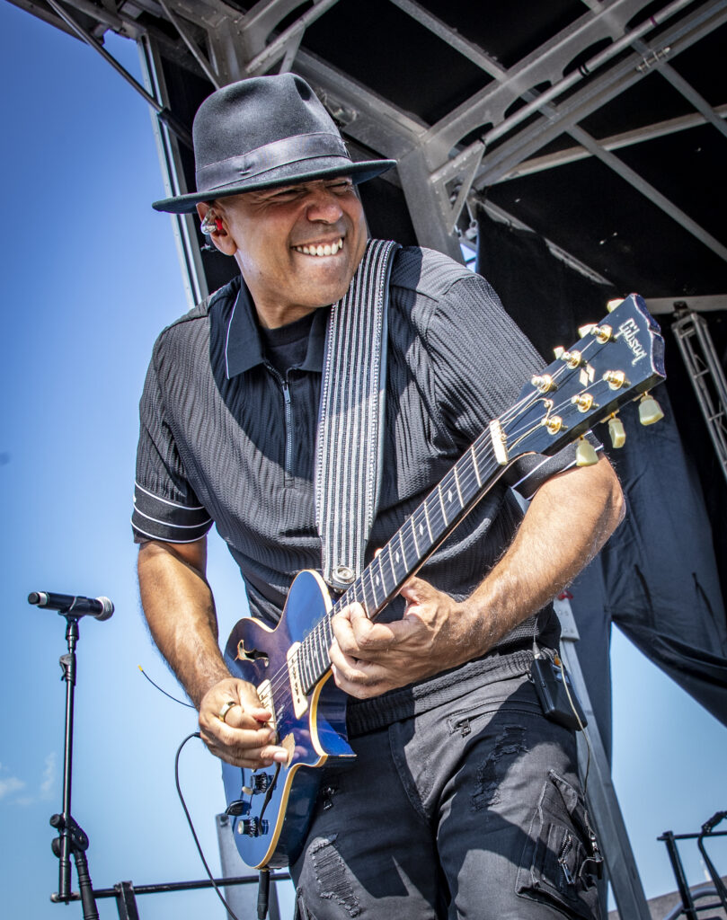 Wayne Baker Brooks performing on stage, playing a blue electric guitar while wearing a black hat and black outfit. He smiles with intensity under a clear blue sky, with stage equipment visible in the background.