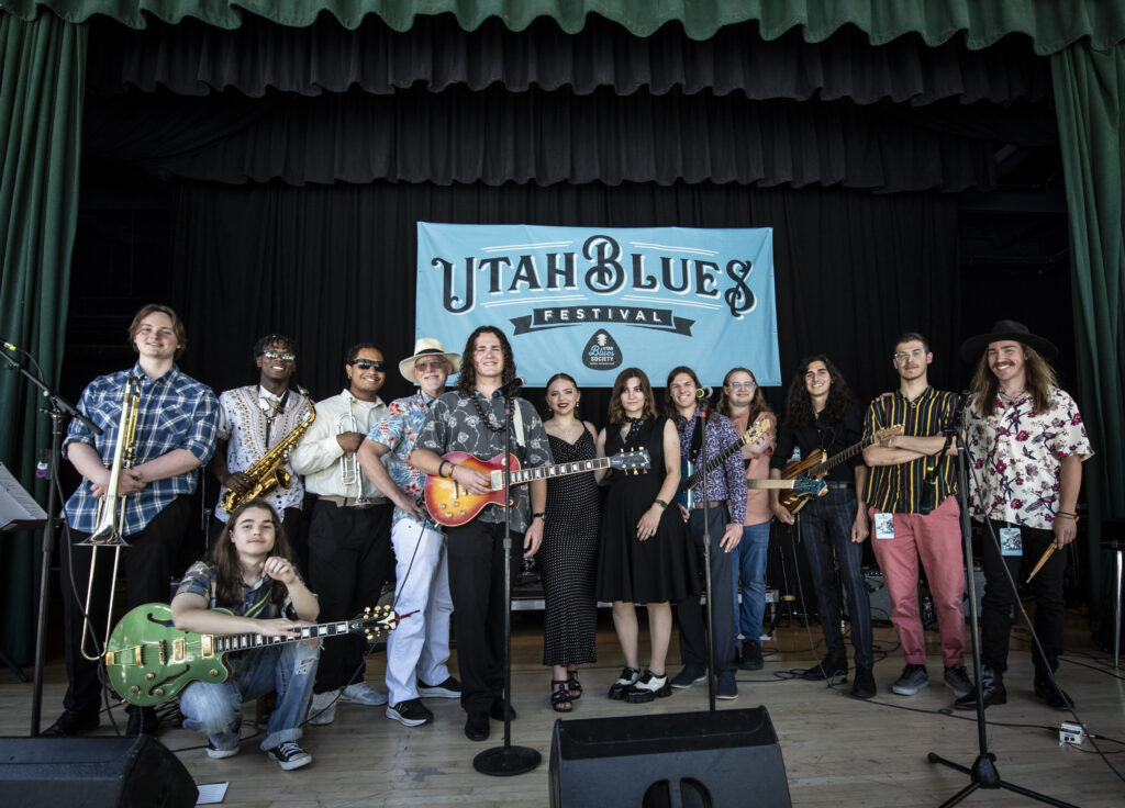 The UBF Youth Blues Band poses together on stage at the Utah Blues Festival. The group consists of young musicians holding various instruments, including guitars, saxophones, a trombone, and drumsticks, smiling under the festival banner. The stage features a vibrant and energetic atmosphere, with green curtains framing the backdrop.