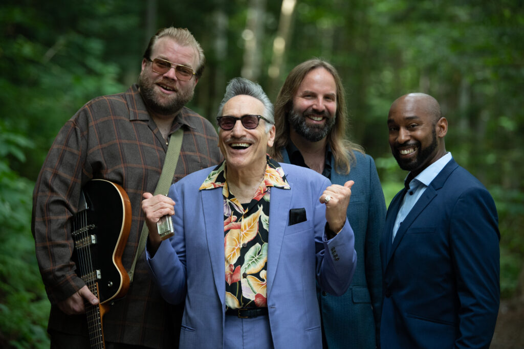 Rick Estrin & The Nightcats band members pose together outdoors in a forested setting. Rick Estrin, in a light purple suit and floral shirt, smiles broadly while holding a harmonica, surrounded by three other band members—one holding a guitar. The group exudes a lively and charismatic energy.